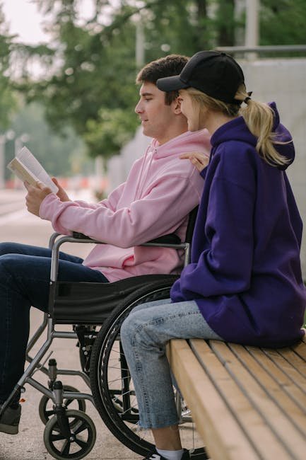 A young man in a wheelchair reading a book with a friend outdoors on a sunny day.